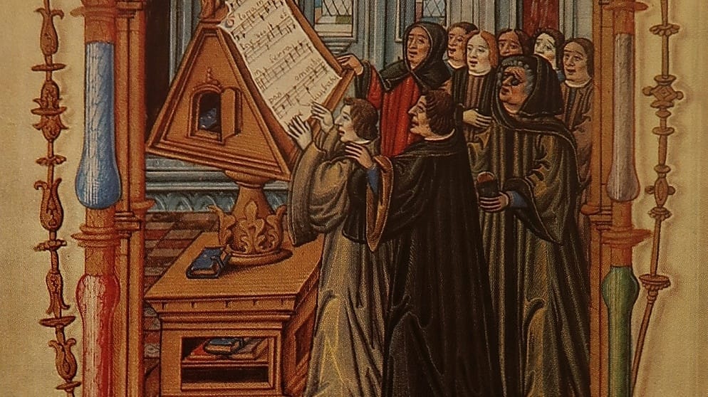 Imagen clásica de Ockeghem donde se le ve dirigiendo un ensaño frente a un gran libro de partituras, con el pelo gris bajo una capucha y unos anteojos negros.